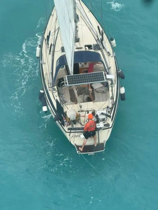 Vista aérea de um veleiro branco em mar aberto, com um socorrista de capacete laranja posicionado na popa para auxiliar os ocupantes no convés. A embarcação apresenta painéis solares e defensas laterais, navegando sobre águas em tons de azul-turquesa sob a luz do dia.