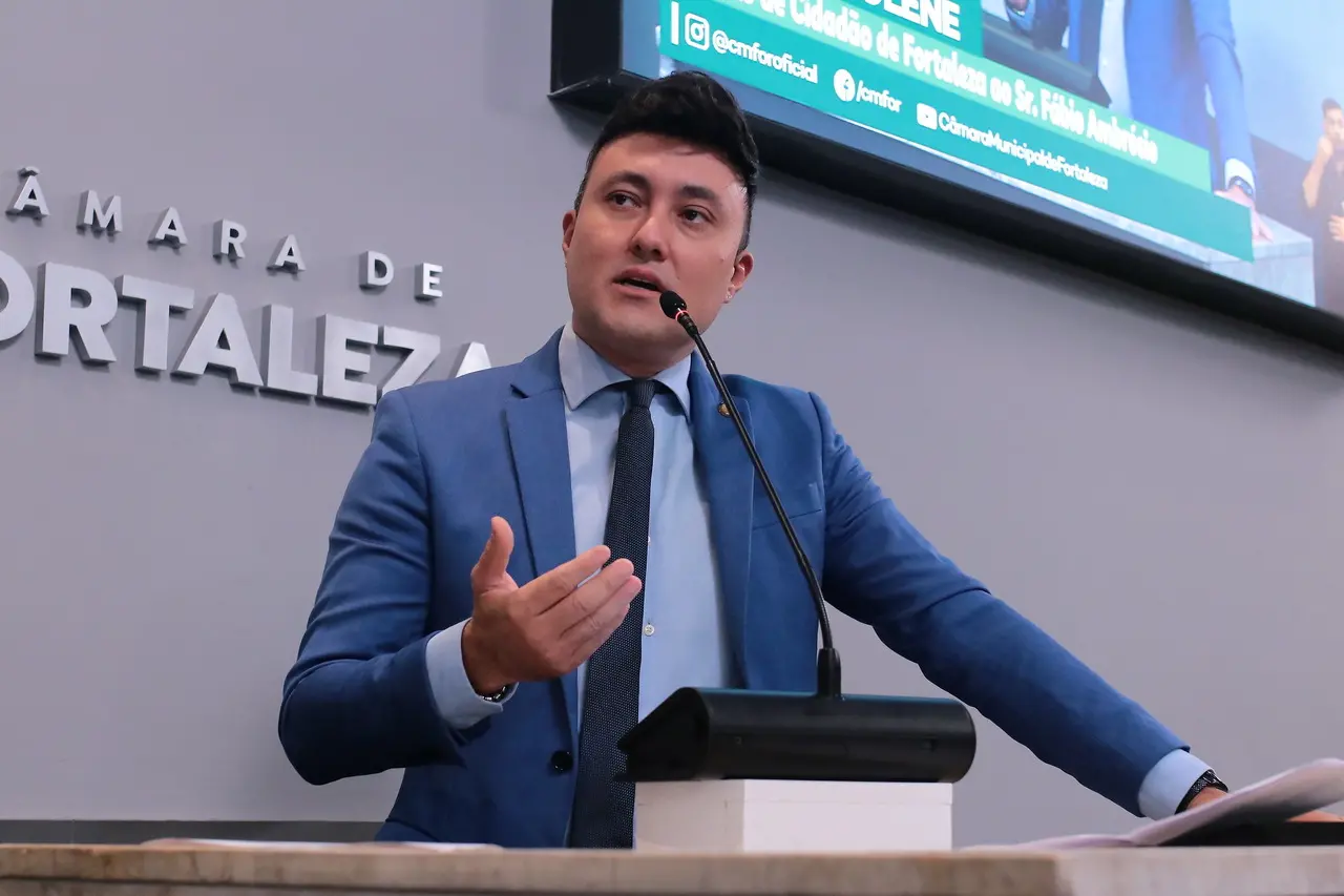 Homem branco de cabelo curto e preto com terno azul e gravata preta discursando em palanque de plenário.