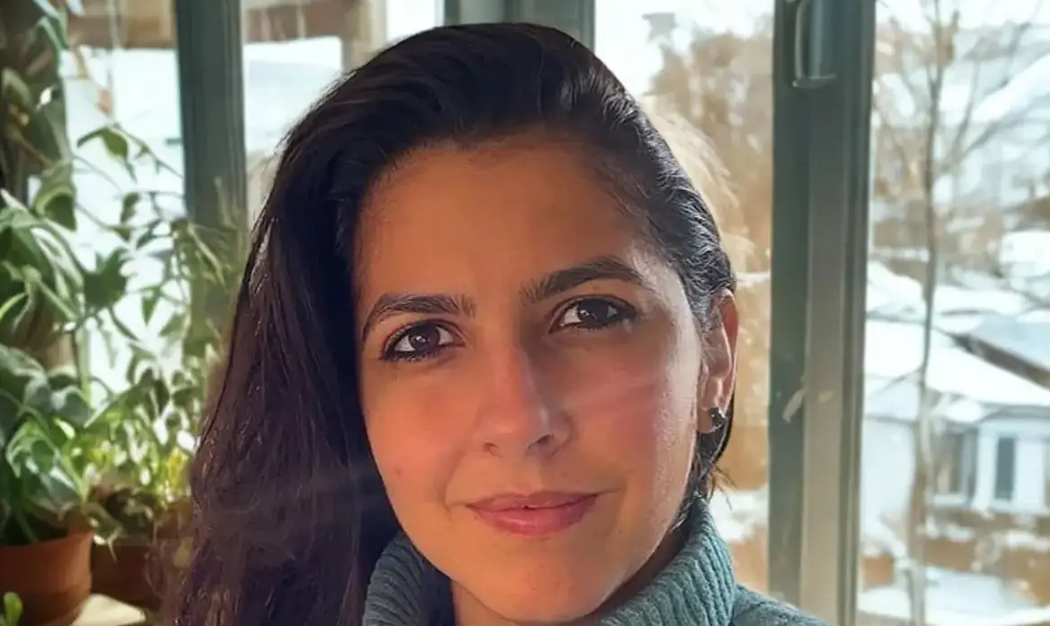 Uma pessoa com cabelo escuro e comprido aparece em um ambiente interno iluminado por luz natural. Ao fundo, há janelas amplas que mostram uma paisagem externa com neve acumulada. Dentro do ambiente, vasos com plantas verdes estão posicionados próximos às janelas. A pessoa veste uma peça de roupa de tom escuro e está enquadrada da altura dos ombros para cima.Forneça seus comentários sobre o BizChat