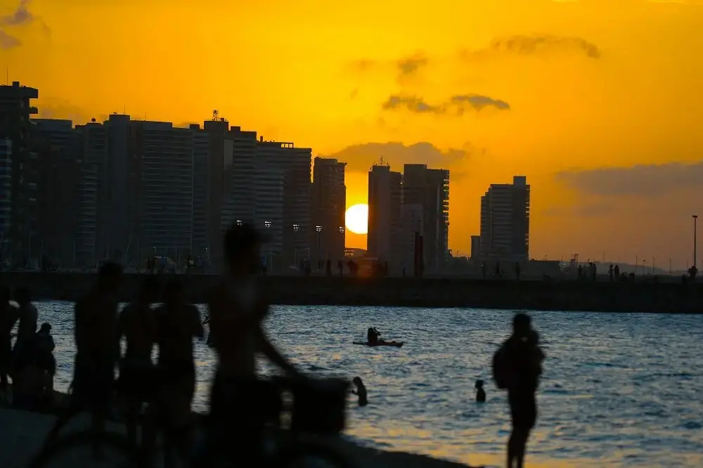 Imagem de pôr do sol na orla de uma cidade, com silhuetas de pessoas e edifícios ao fundo, refletindo na água. Ideal para quem busca fotos de sunset urbano.