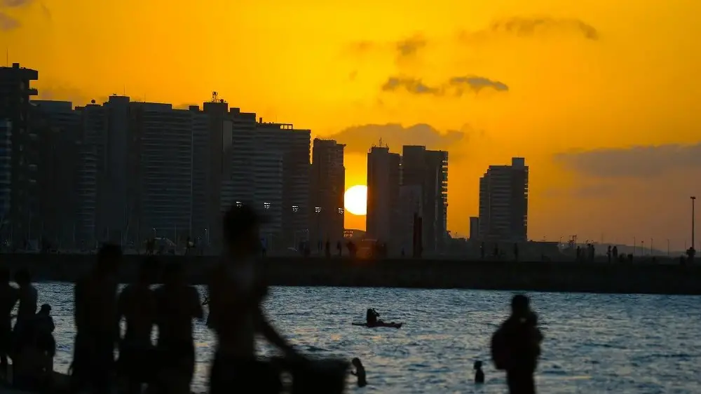 Imagem de pôr do sol na orla de uma cidade, com silhuetas de pessoas e edifícios ao fundo, refletindo na água. Ideal para quem busca fotos de sunset urbano.