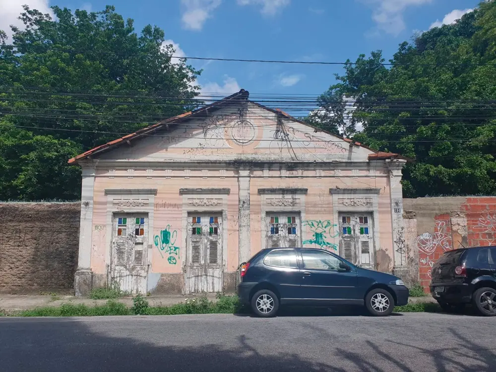 Na imagem, fachada de uma casa histórica de um pavimento no bairro da Parangaba, em Fortaleza. A construção possui paredes em tom rosa desbotado com detalhes arquitetônicos clássicos e um frontão triangular central ornamentado. Há quatro portas/janelas altas de madeira branca com vidros coloridos na parte superior, todas apresentando sinais de desgaste e pichações. À frente da casa, um carro azul escuro está estacionado na rua asfáltica. Ao fundo, árvores verdes sob um céu azul com nuvens brancas.