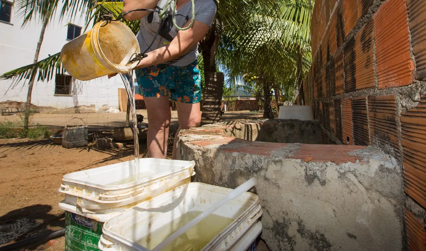 Mulher de blusa e short despeja água de um balde em recipientes plásticos sobre um poço artesanal de tijolos, em área com coqueiros ao fundo.