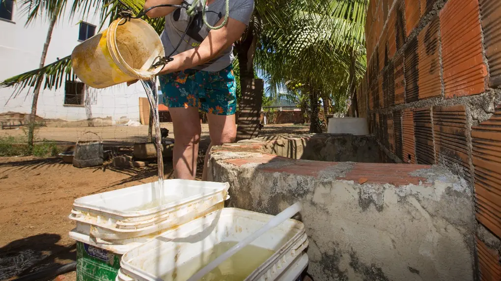 Mulher de blusa e short despeja água de um balde em recipientes plásticos sobre um poço artesanal de tijolos, em área com coqueiros ao fundo.