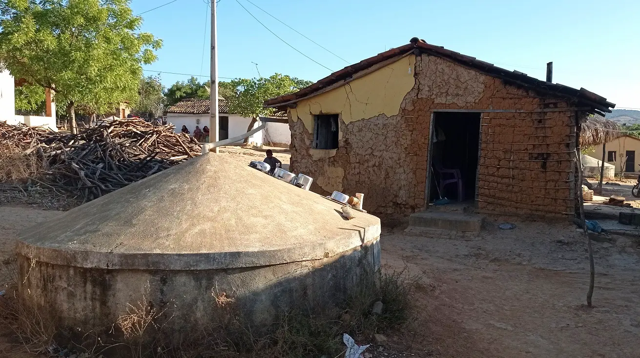Cisterna de concreto em frente a uma casa de taipa rústica no Crato, com lenha empilhada e céu limpo ao fundo.