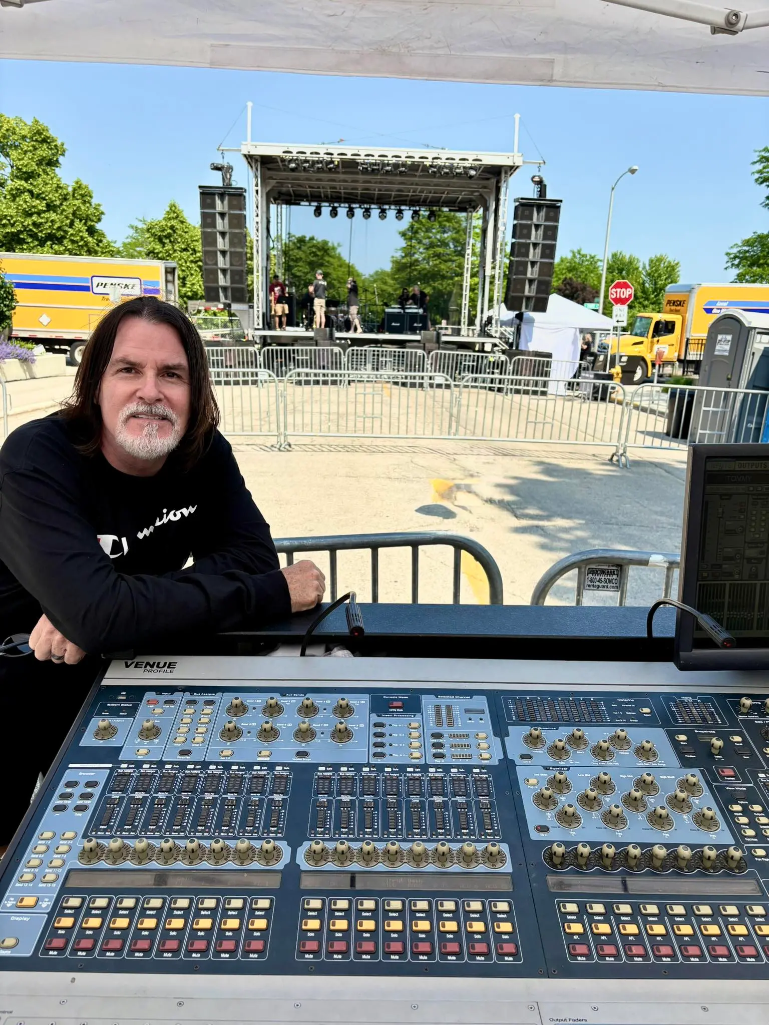 Tommy DeCarlo posa para uma foto diante do palco de um show. Ele está sentado e com os braços encostados em uma mesa de som.