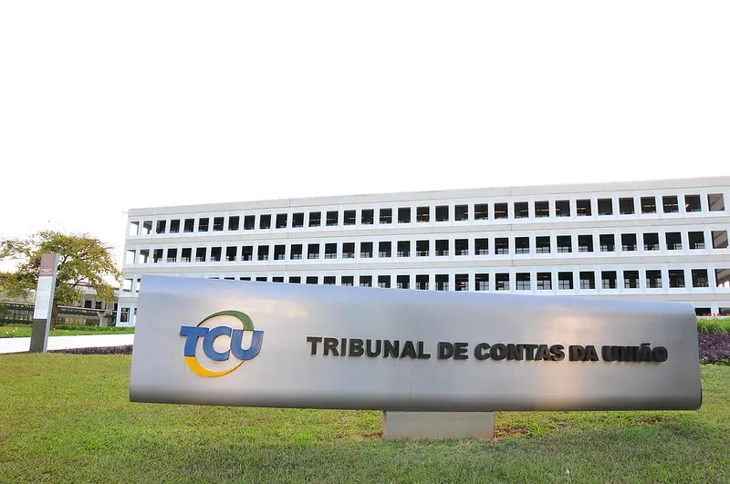 Fachada da sede do Tribunal de Contas da União (TCU) em Brasília. Em primeiro plano, um totem cinza exibe o logotipo colorido do TCU e o nome da instituição por extenso. Ao fundo, vê-se o edifício principal de arquitetura moderna com janelas retangulares repetitivas sob um céu claro.