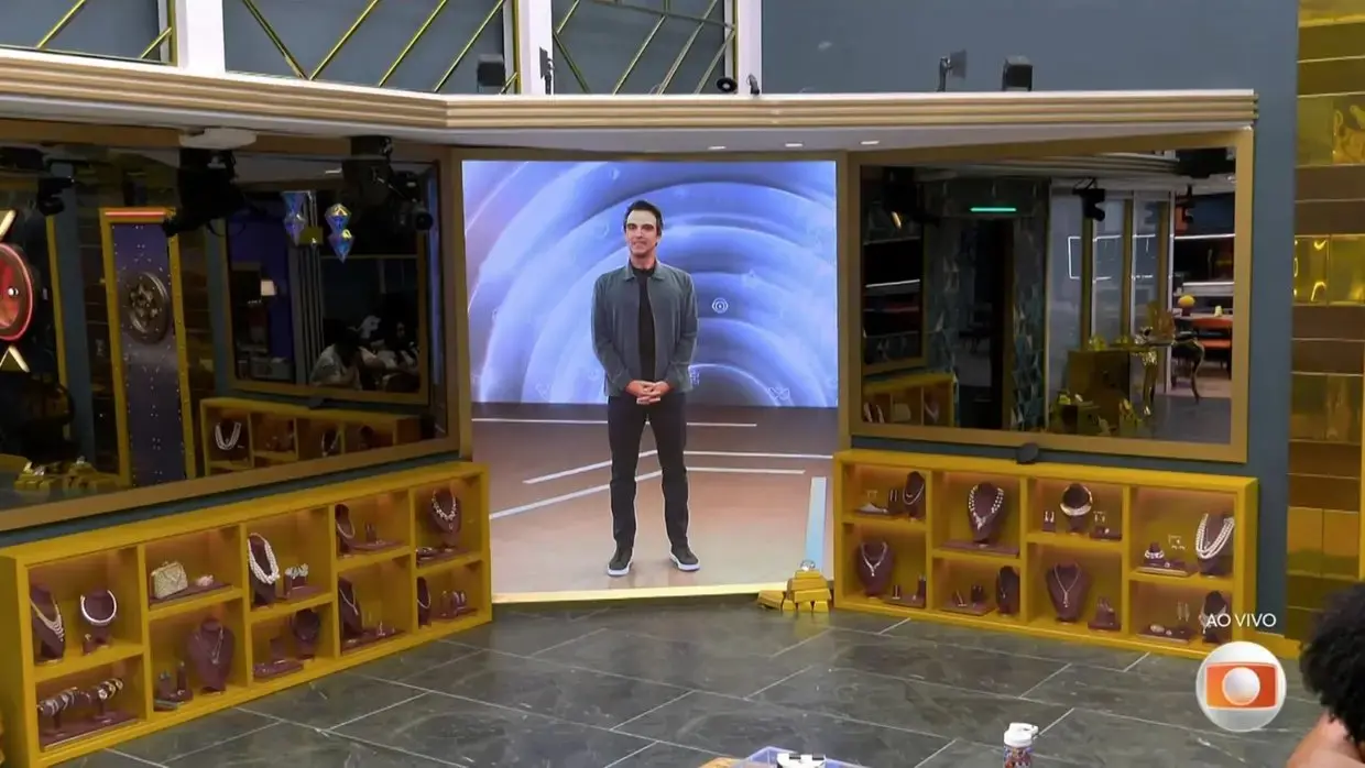 Tadeu no telão da casa do BBB. Ele aparece de calça presta, camiseta preta e uma jaqueta cinza. O apresentador está de pé no estúdio.