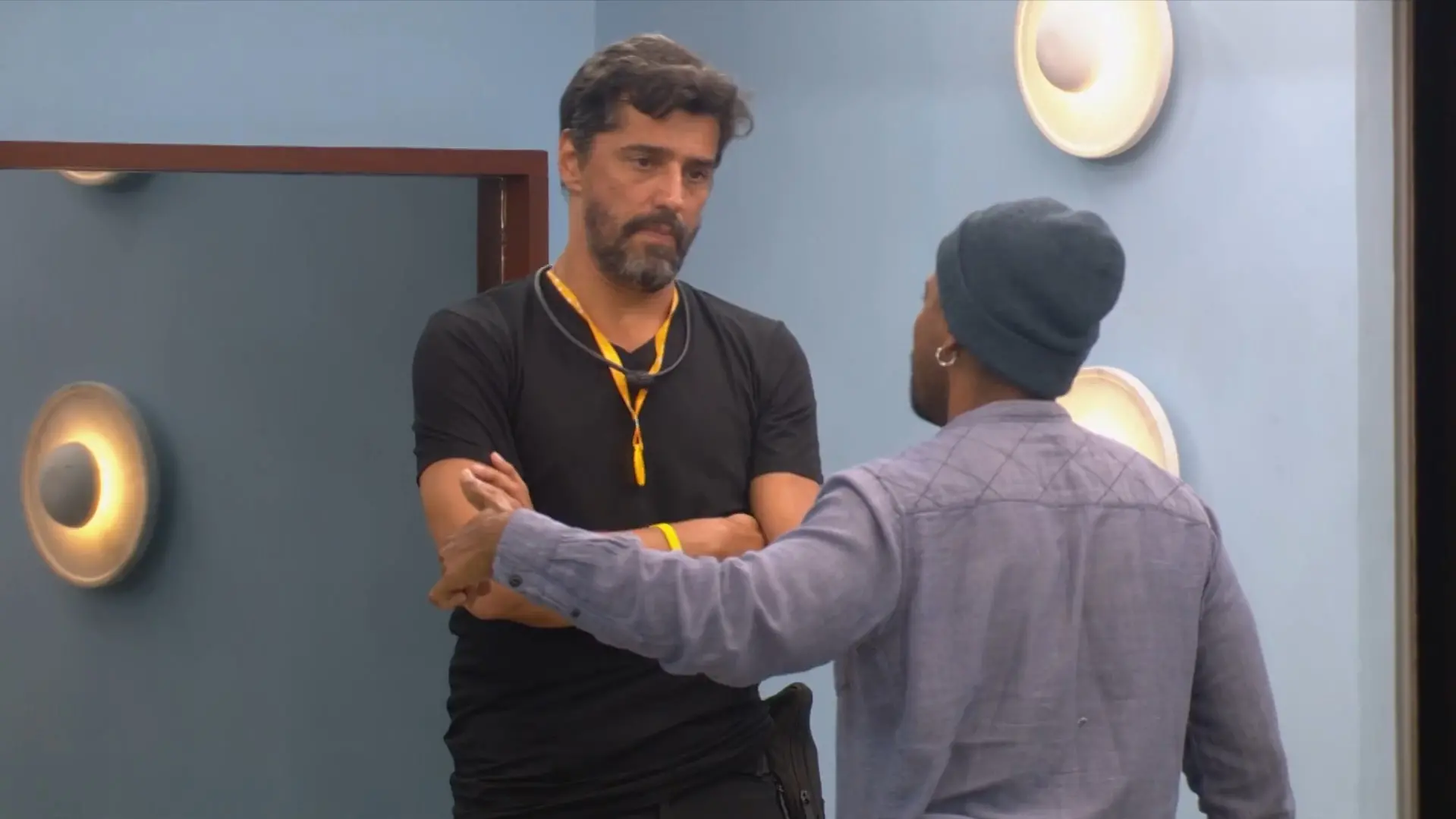 Alberto Cowboy, de braços cruzados e expressão séria, encara Leandro Boneco enquanto este gesticula durante uma conversa em um ambiente de paredes azuis. A cena captura um momento de tensão entre os dois homens, iluminada por luminárias circulares fixadas na parede ao fundo.