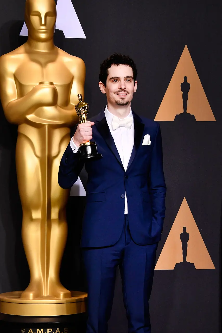 O diretor Damien Chazelle posa segurando a estatueta do Oscar, vestindo um terno azul-marinho com lapelas pretas e gravata borboleta branca. Ao fundo, destaca-se o logotipo dourado da premiação sobre uma parede escura, compondo uma cena solene e vitoriosa.