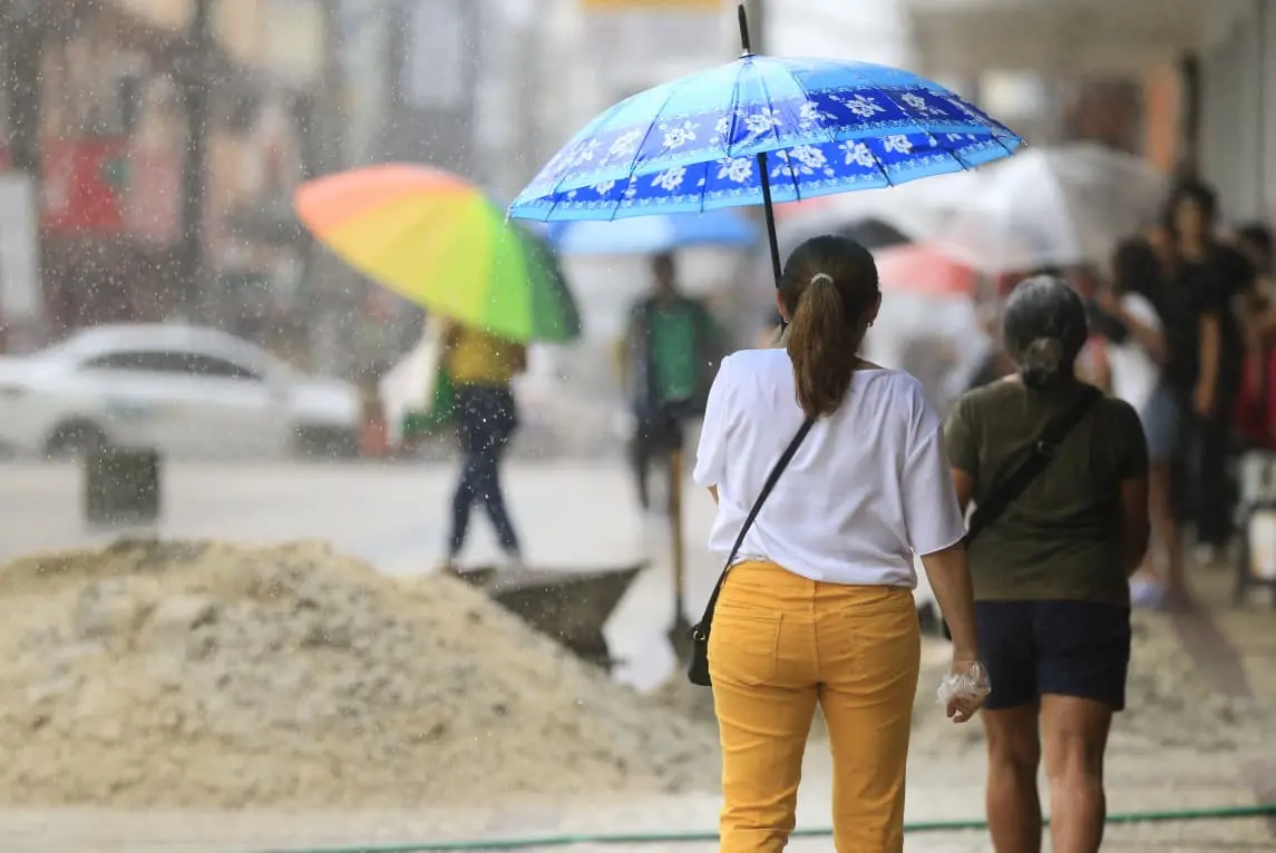 Pessoas caminham sob chuva em calçada urbana. Em destaque, uma mulher de calça amarela segura um guarda-chuva azul. Ao fundo, pedestres, um guarda-chuva colorido e montes de areia de uma obra.
