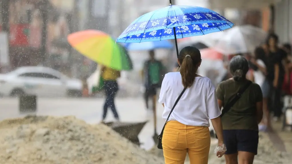 Pessoas caminham sob chuva em calçada urbana. Em destaque, uma mulher de calça amarela segura um guarda-chuva azul. Ao fundo, pedestres, um guarda-chuva colorido e montes de areia de uma obra.