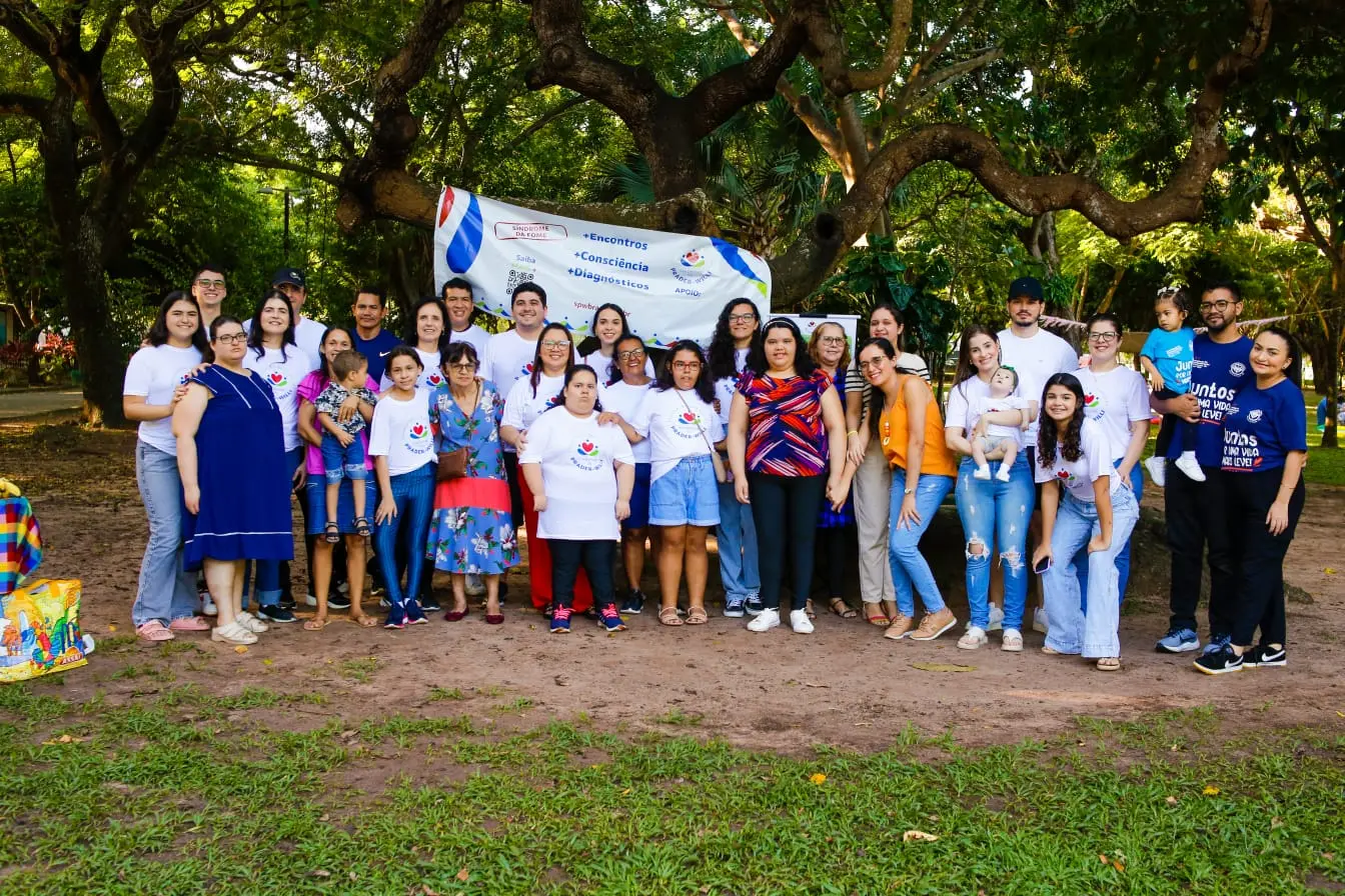 Grupo de famílias e crianças reunido em um parque em Fortaleza, sob árvores, com faixa sobre Prader-Willi ao fundo.