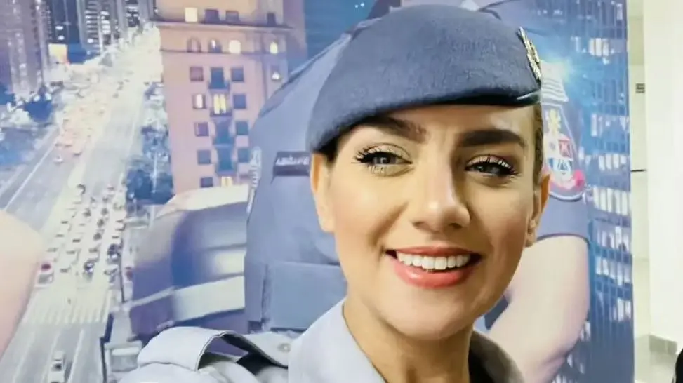 Uma pessoa vestindo uniforme policial aparece em primeiro plano dentro de um ambiente interno. Ao fundo, há um grande painel com a imagem de uma cidade iluminada à noite e a foto ampliada de uma pessoa também usando uniforme policial.