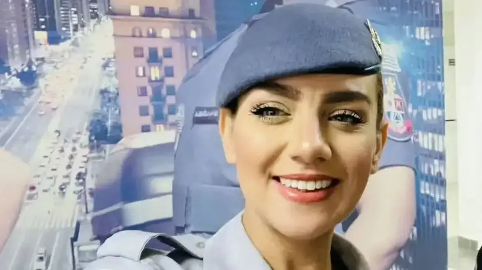 PM morta no apartamento com tiro na cabeça vestindo uniforme aparece em primeiro plano dentro de um ambiente interno. Ao fundo, há um grande painel com a imagem de uma cidade iluminada à noite e a foto ampliada de uma pessoa também usando uniforme policial. Imagem usada em matéria sobre policiais femininas supostamente ido ao imóvel para limpar.