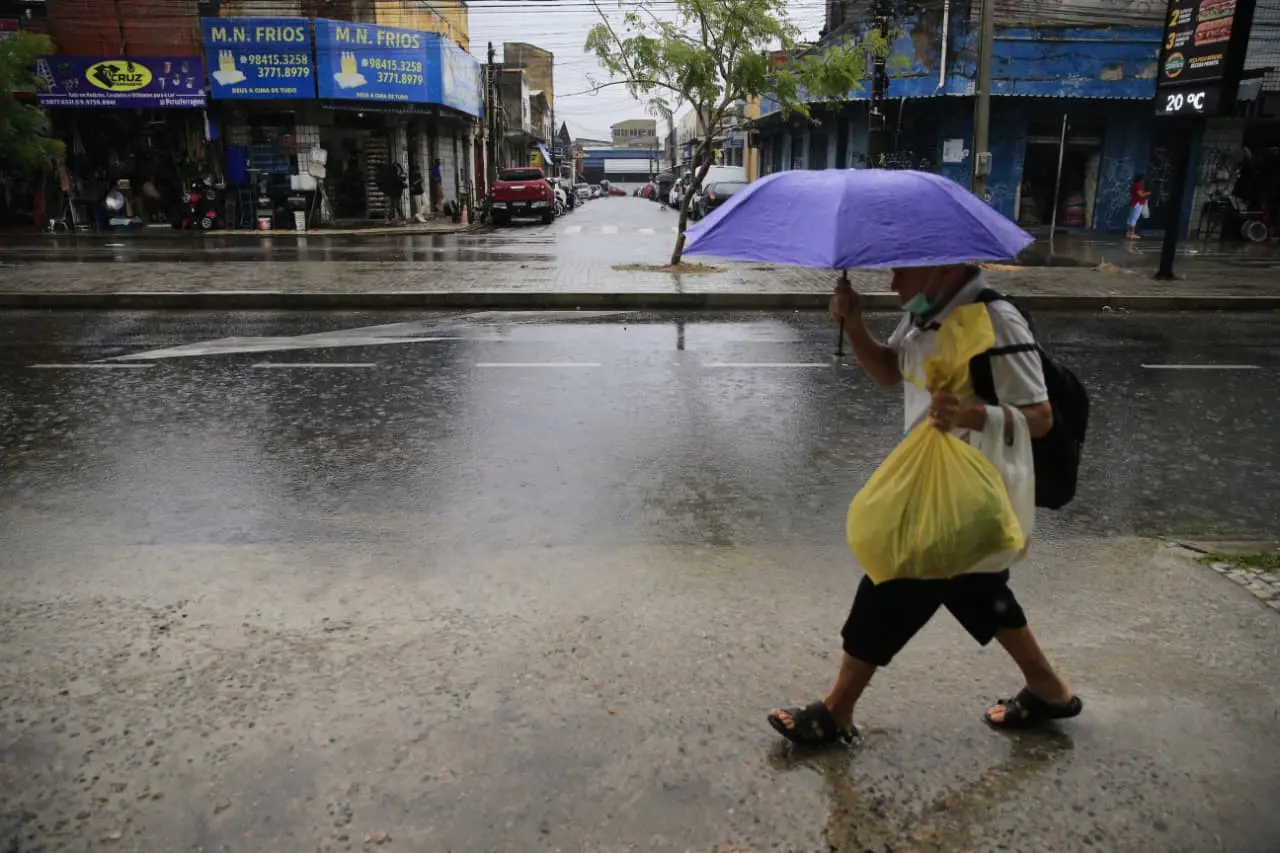 Homem caminha sob chuva em rua urbana, segurando guarda-chuva roxo e sacolas amarelas. Ao fundo, comércios com fachadas azuis e calçada molhada. Registro do cotidiano na cidade.