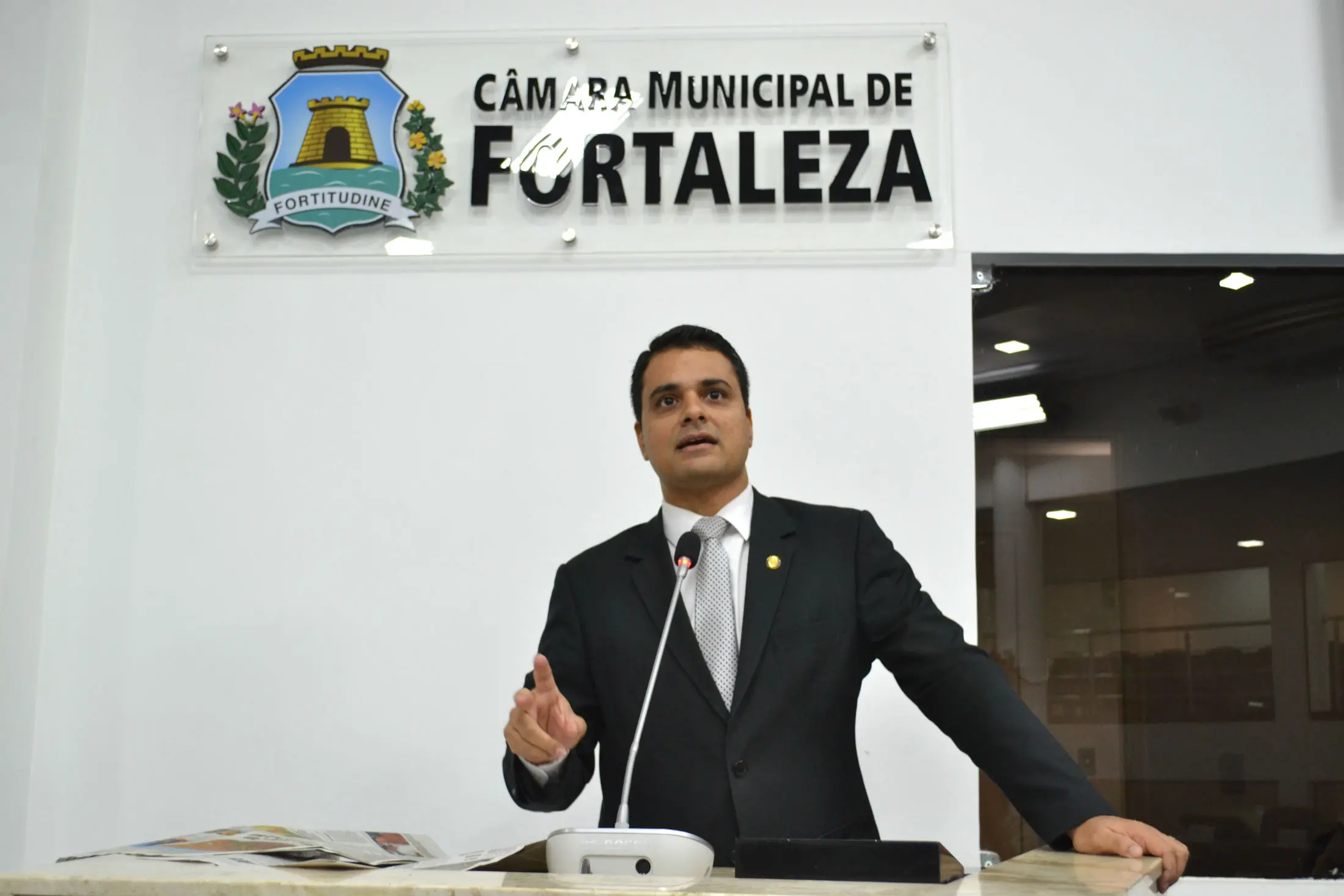 Homem de terno falando em um púlpito com microfone na Câmara Municipal de Fortaleza, com o brasão da cidade e a placa “Câmara Municipal de Fortaleza” na parede ao fundo. Trata-se de Gardel Rolim.