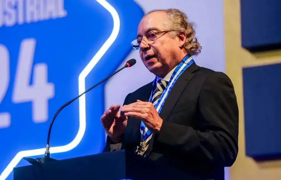 Mário Araripe é um homem idoso de cabelos grisalhos e que usa óculos de grau. Na foto, ele usa terno preto e está discursando no palco.