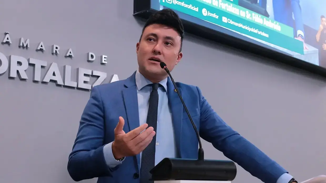 Homem branco de cabelo curto e preto com terno azul e gravata preta discursando em palanque de plenário.