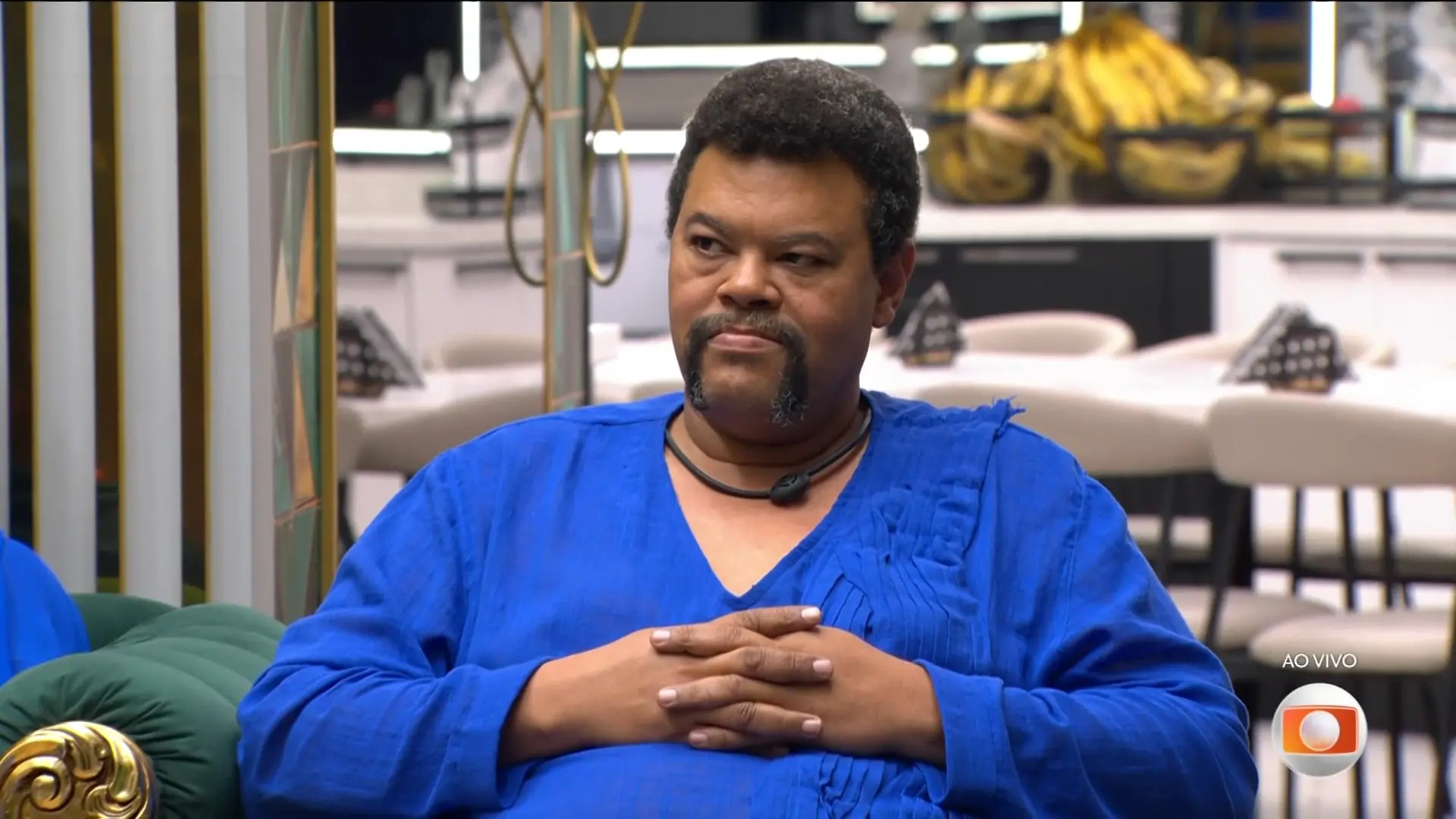 Uma pessoa sentada em um sofá verde com detalhes dourados, vestindo uma roupa azul vibrante de mangas longas. As mãos estão apoiadas sobre o colo, com os dedos entrelaçados. Ao fundo, há um ambiente interno bem iluminado, com mesas, cadeiras claras e uma bancada onde aparecem recipientes com bananas. No canto inferior direito da imagem, há um logotipo de transmissão ao vivo de uma emissora de televisão.