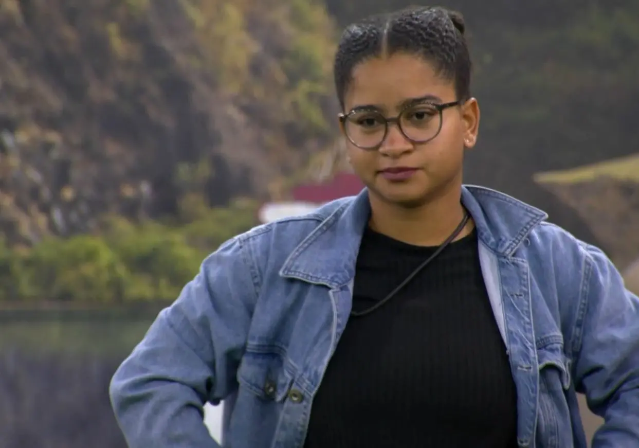 Uma pessoa em ambiente externo posando com os braços apoiados na cintura, vestindo uma jaqueta jeans sobre uma blusa escura. Ao fundo, há uma paisagem composta por um corpo d’água e encostas rochosas com vegetação.