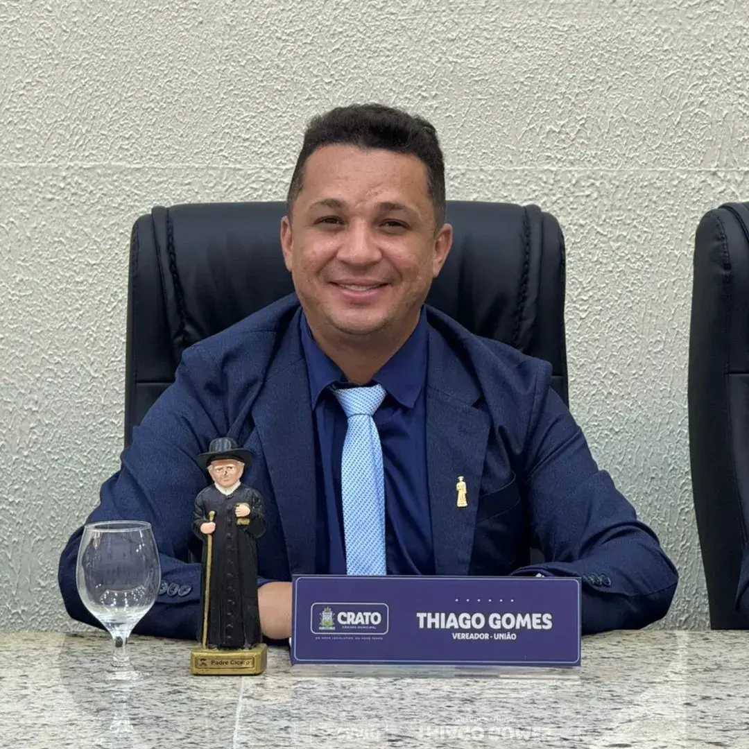 O vereador Thiago Gomes posa em sua bancada na Câmara do Crato. Ele tem uma taça, uma estátua de Padre Cícero e uma placa com seu nome.