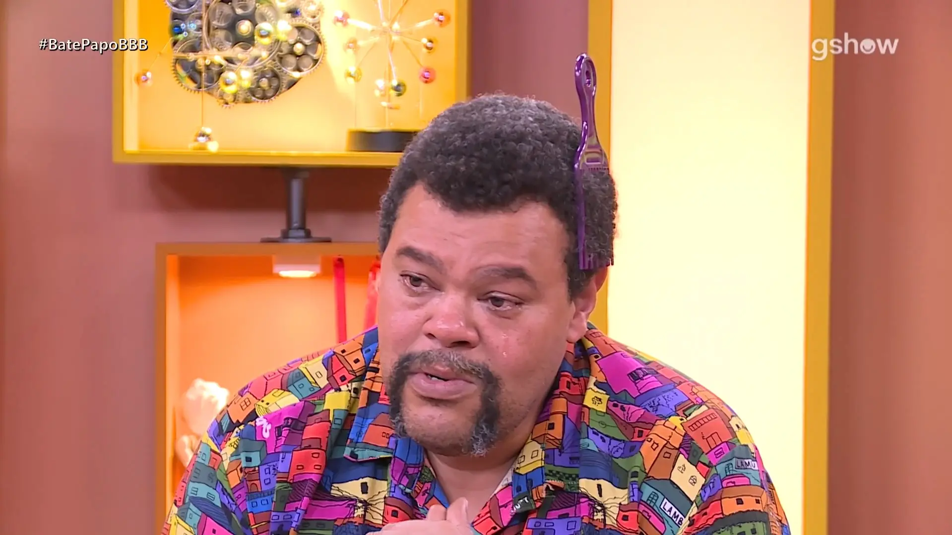 O ator Babu Santana aparece emocionado e com lágrimas nos olhos, vestindo uma camisa colorida com estampas de casas coloridas e um pente garfo roxo espetado no cabelo black power. Ele está em um cenário de estúdio com tons de amarelo e laranja, destacando sua expressão sensível durante uma entrevista após ser eliminado do BBB 26.