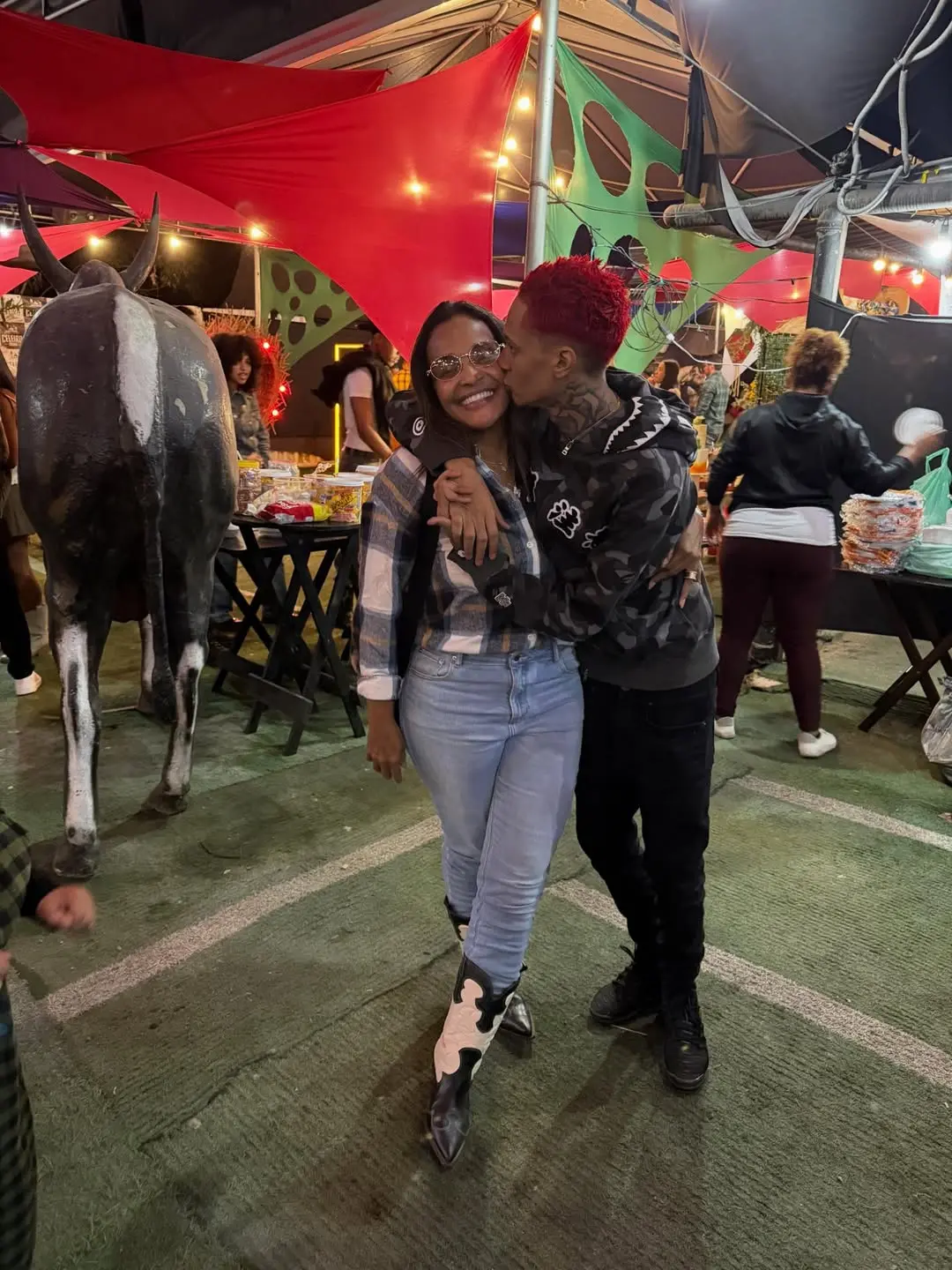 Márcia Gama, mãe do rapper Oruam, e o artista posam abraçados em um evento festivo decorado com tendas coloridas e luzes. Eles posam sorridentes em um ambiente com chão de grama sintética, onde se nota a presença de uma escultura de boi ao lado e mesas de petiscos ao fundo.