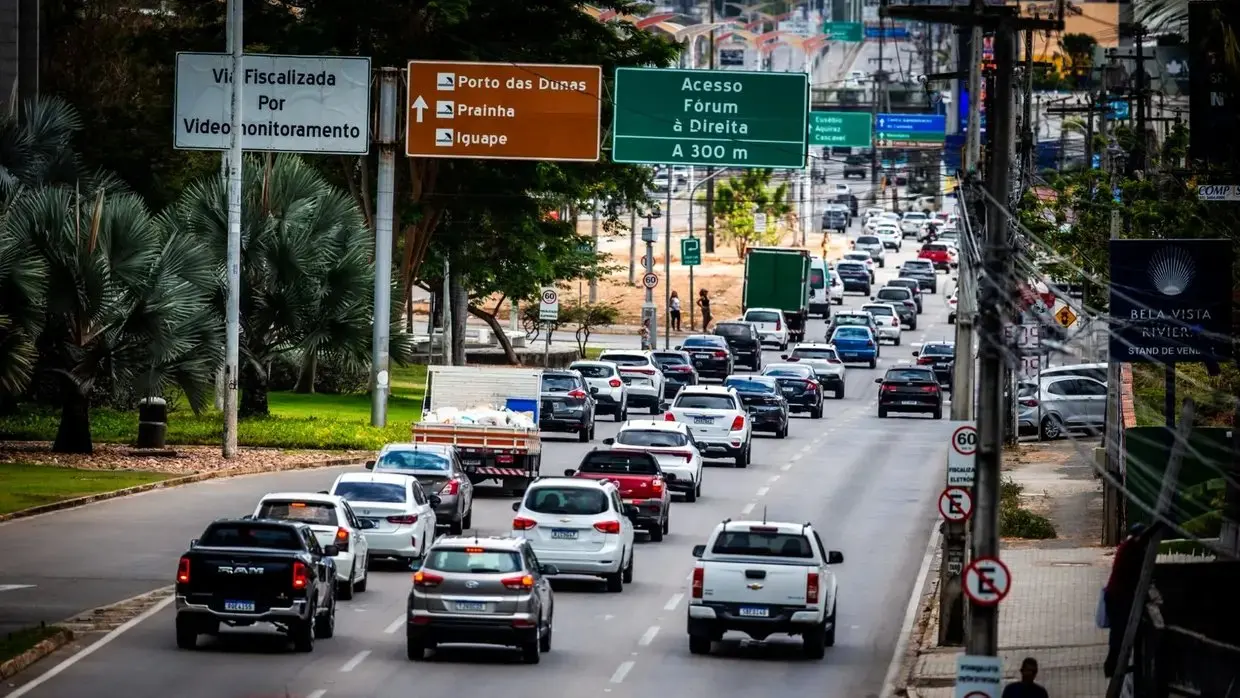 Trânsito intenso em uma avenida de cidade brasileira, com vários carros e placas de sinalização lado a lado, indicando acessos e destinos na cidade.