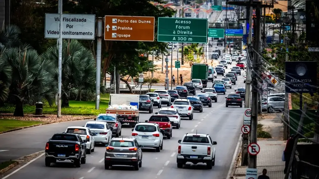 Trânsito intenso em uma avenida de cidade brasileira, com vários carros e placas de sinalização lado a lado, indicando acessos e destinos na cidade.