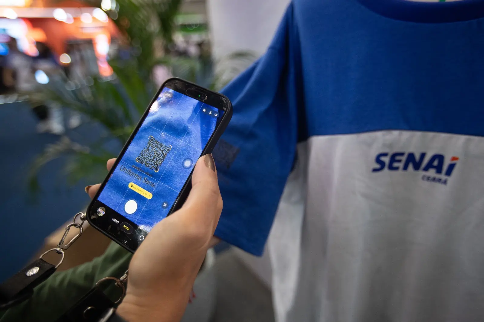 Uma mão segura um smartphone que aponta a câmera para um código QR estampado na manga azul de uma camiseta branca com o logotipo 