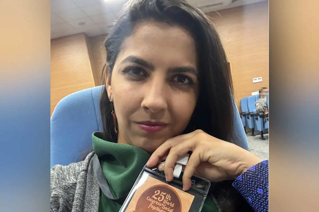 Uma pessoa está sentada em uma poltrona acolchoada de cor azul dentro de um auditório iluminado, com paredes revestidas em madeira clara. A pessoa segura um crachá próximo ao peito; o crachá tem fundo alaranjado e exibe o texto “25th World Congress of Social Psychiatry”. Ao fundo, há outras poltronas semelhantes e detalhes arquitetônicos do ambiente.