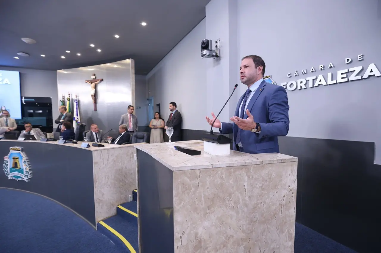Homem branco de cabelo curto castanho escuro com terno azul em cima de tribuna de plenário, discursando no microfone.