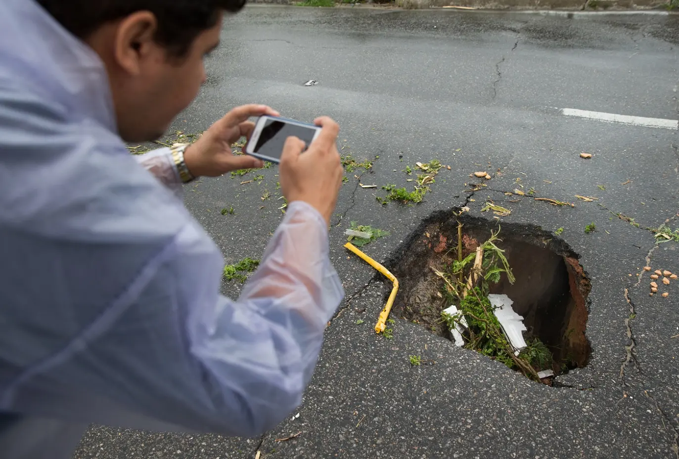 Homem de capa de chuva fotografando um grande buraco em estrada de asfalto molhada com seu smartphone, em Fortaleza.