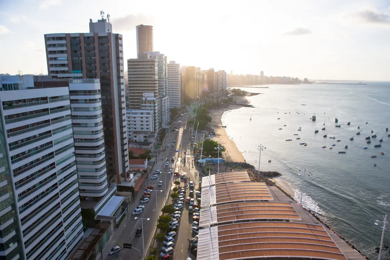 Vista aérea da orla de Fortaleza com prédios altos à beira-mar, avenida com carros em circulação e faixa de praia com pequenas embarcações no mar, sob luz do fim de tarde.