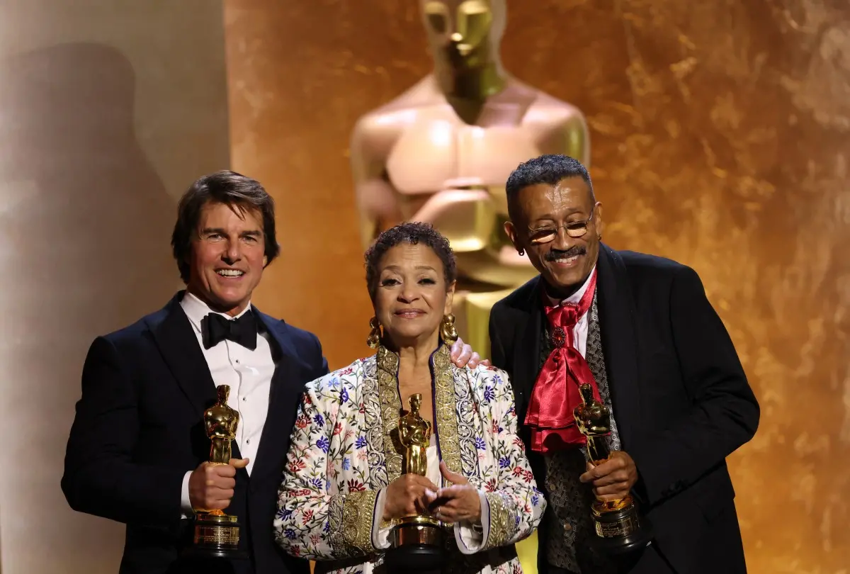 Três pessoas no palco celebrando após receber o prêmio Oscar, com um fundo dourado e uma estatueta do Oscar em mãos, símbolo do cinema de destaque.