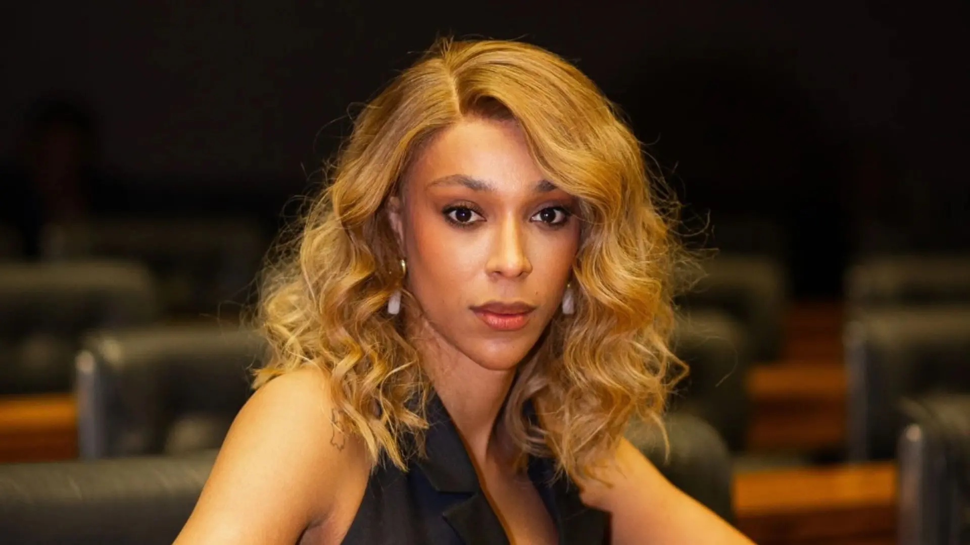 Mulher com cabelos cacheados e loiros, looks elegante, sentada em ambiente profissional, com expressão confiante, ideal para conteúdo sobre beleza e estilo.