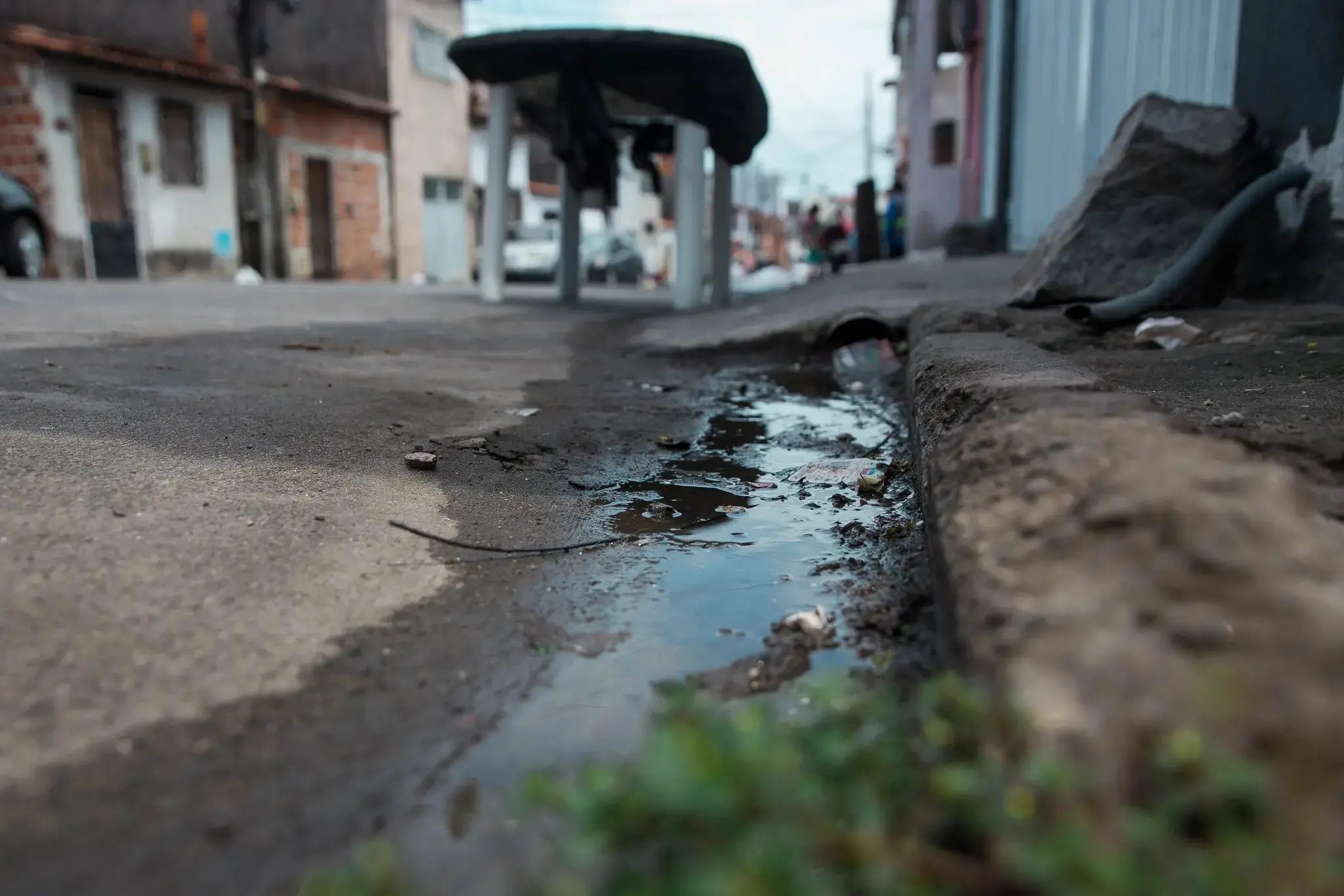 Cenário urbano com uma sarjeta suja e água empoçada escorrendo, com lixo visível e construções desfocadas ao fundo.