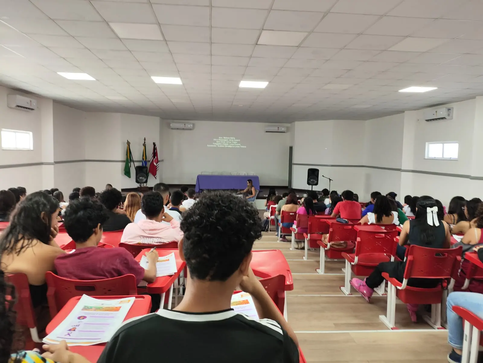 Auditório lotado com estudantes sentados em carteiras vermelhas, de costas para a câmera, assistindo a um palestrante no palco, com um projetor exibindo texto ao fundo.