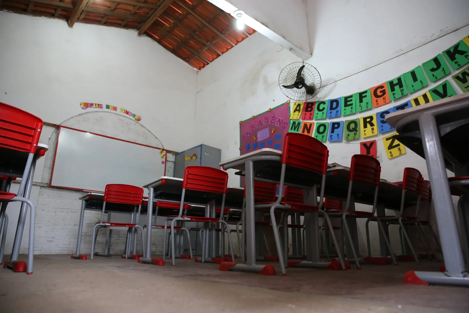 Uma sala de aula vazia com carteiras e cadeiras vermelhas, lousa branca e um alfabeto colorido decorando a parede.