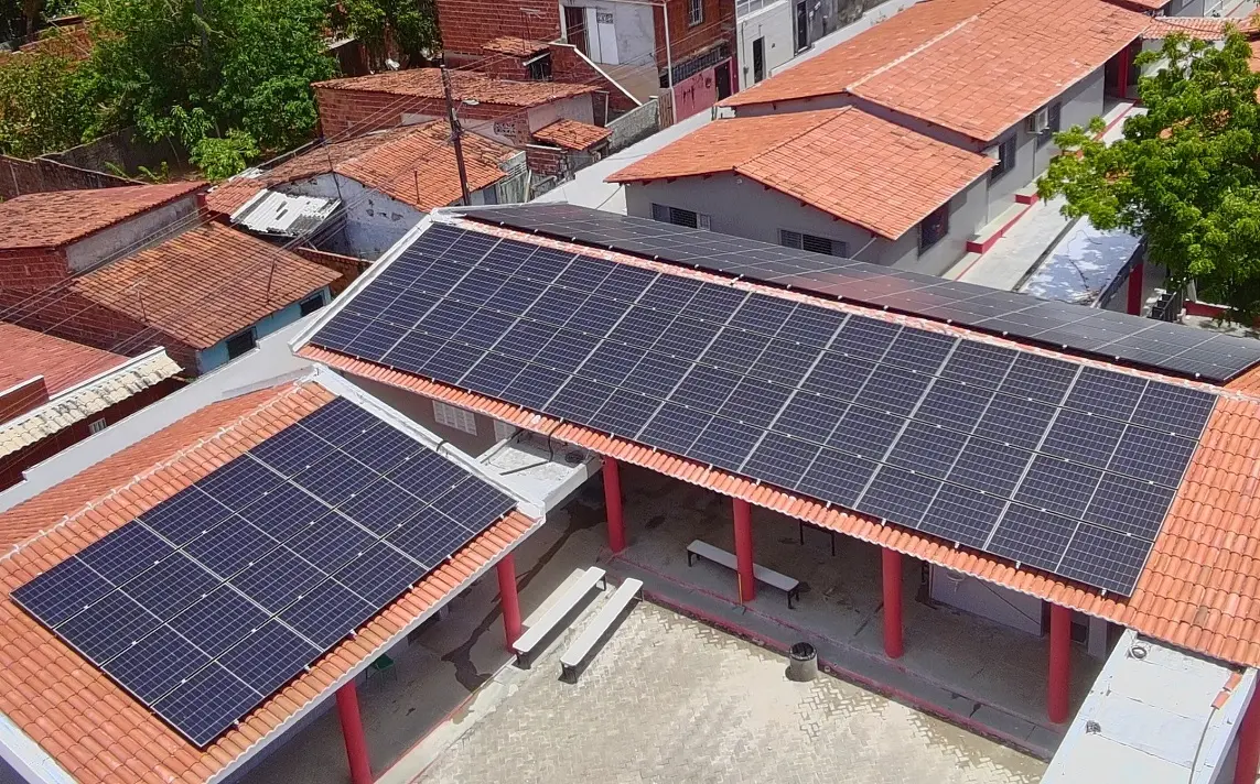 Vista aérea de um complexo escolar com telhados de telha colonial e grandes instalações de painéis solares fotovoltaicos, em um ambiente urbano de Caucaia.