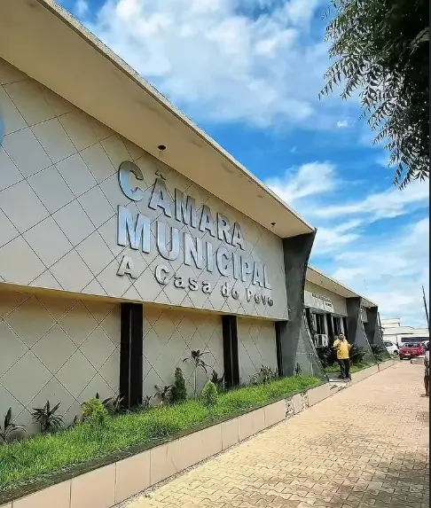 Fachada de um prédio da Câmara Municipal com a inscrição “Câmara Municipal – A Casa do Povo” em letras grandes na parede. O edifício tem arquitetura moderna, com paredes claras e linhas geométricas. Há um caminho de piso intertravado ao lado, com uma pessoa caminhando e carros estacionados ao fundo. O céu está azul com algumas nuvens e há árvores próximas ao prédio.