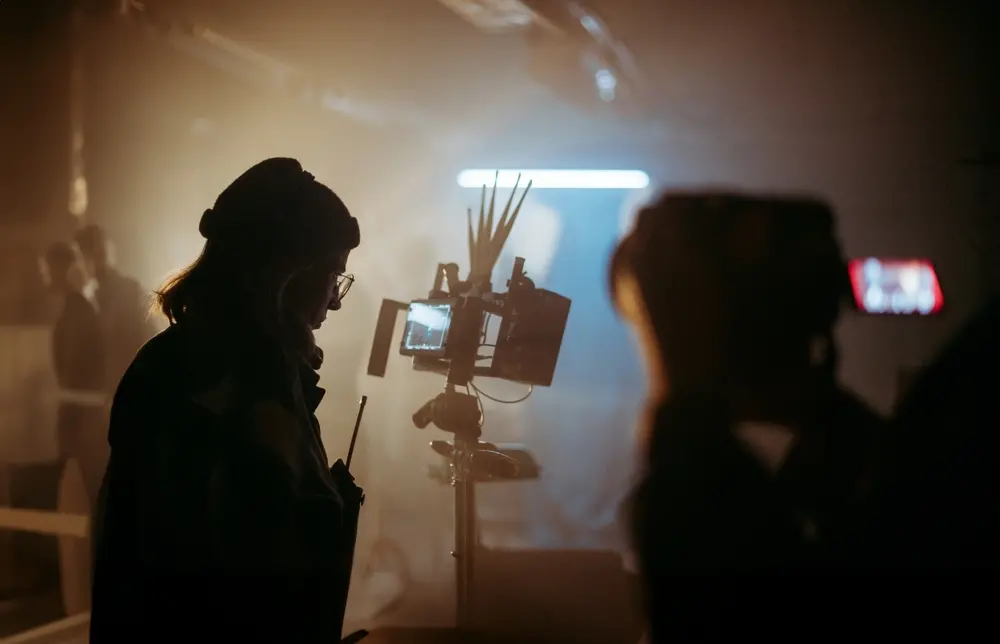 Cena de uma filmagem ou gravação com duas pessoas ao fundo, uma delas ao lado de uma câmera em um ambiente com iluminação suave e atmosfera de estúdio.
