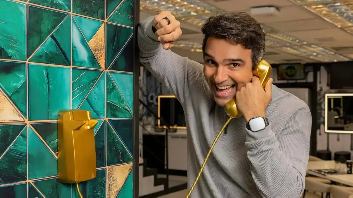 Homem sorridente em escritório usando telefone antigo amarelo com parede decorativa de azulejos verdes ao fundo.