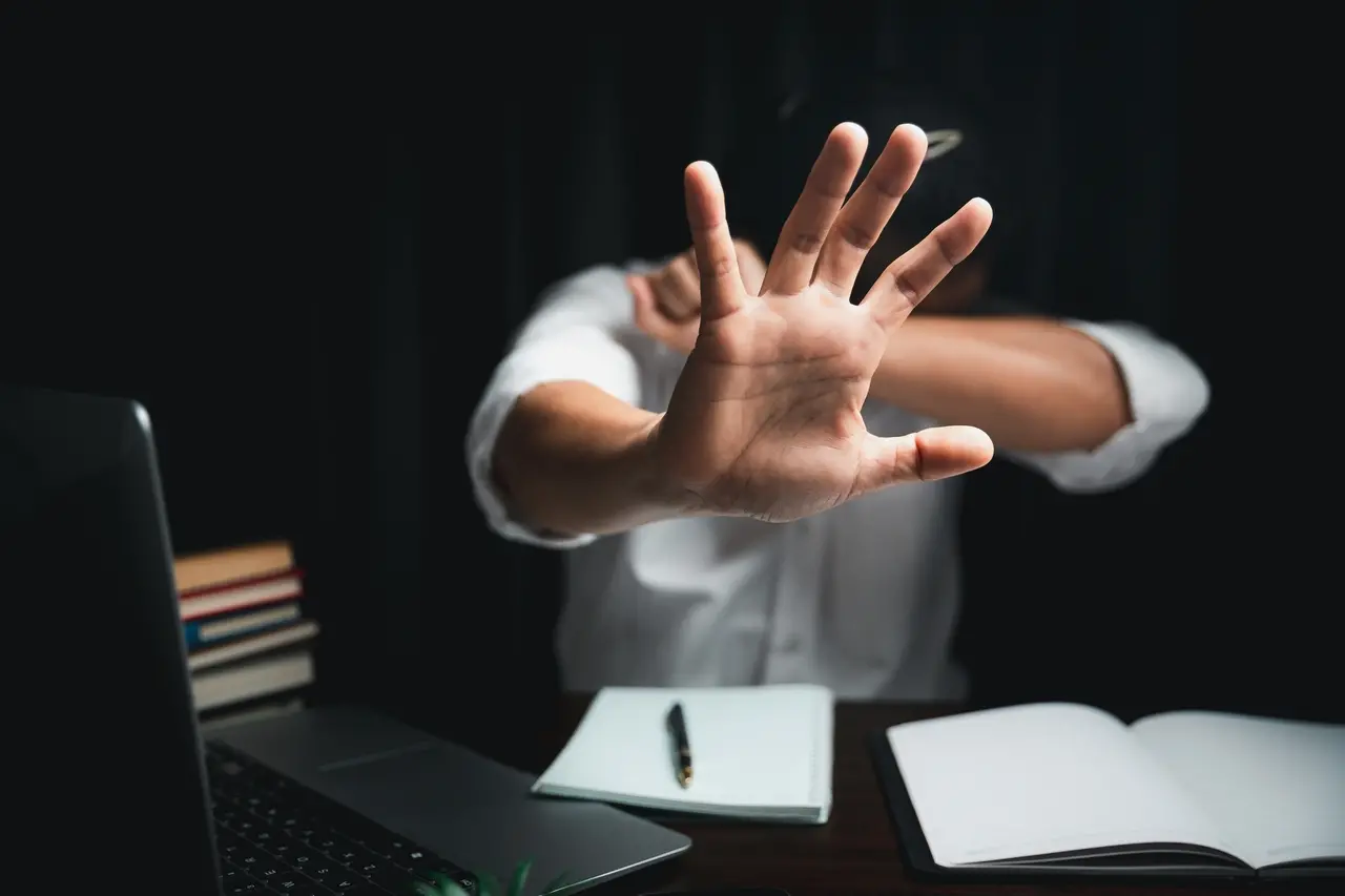 Mulher usando a mão para sinal de 'pare' durante trabalho ou estudo, sentado em frente ao notebook com livros e caderno ao lado.