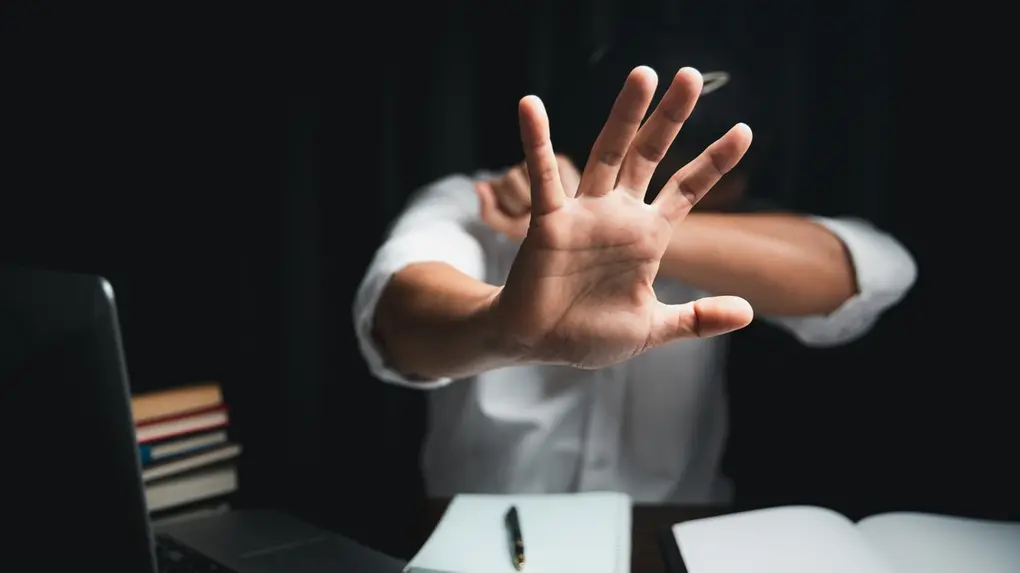 Mulher usando a mão para sinal de 'pare' durante trabalho ou estudo, sentado em frente ao notebook com livros e caderno ao lado.