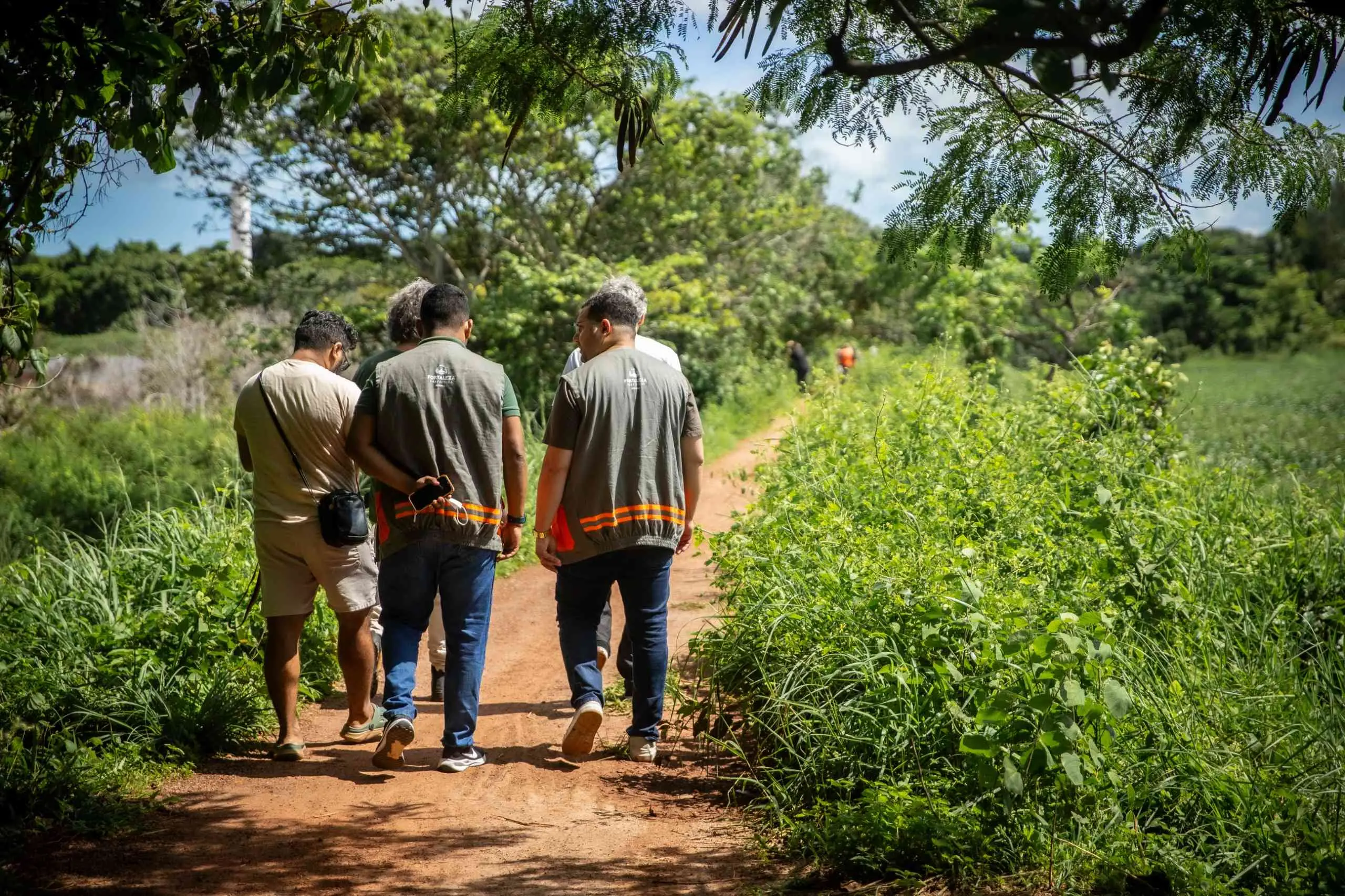 Um grupo de homens caminha por uma trilha de terra em meio a uma vegetação densa e exuberante, alguns vestindo coletes de trabalho com detalhes refletivos.