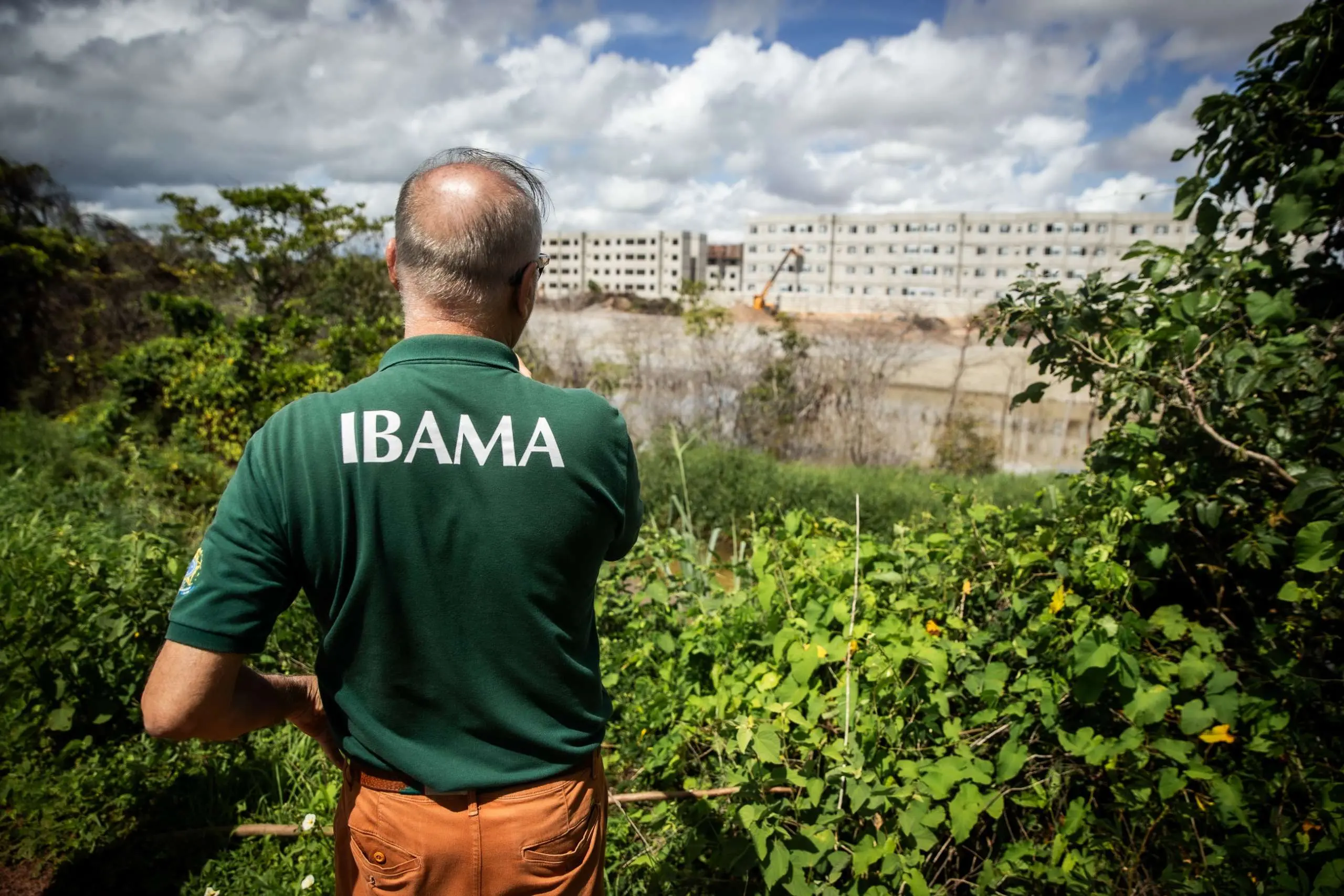 Fiscal com farda verde do IBAMA, de costas, observa obra de prédios residenciais a partir de uma área de preservação ambiental com densa vegetação.