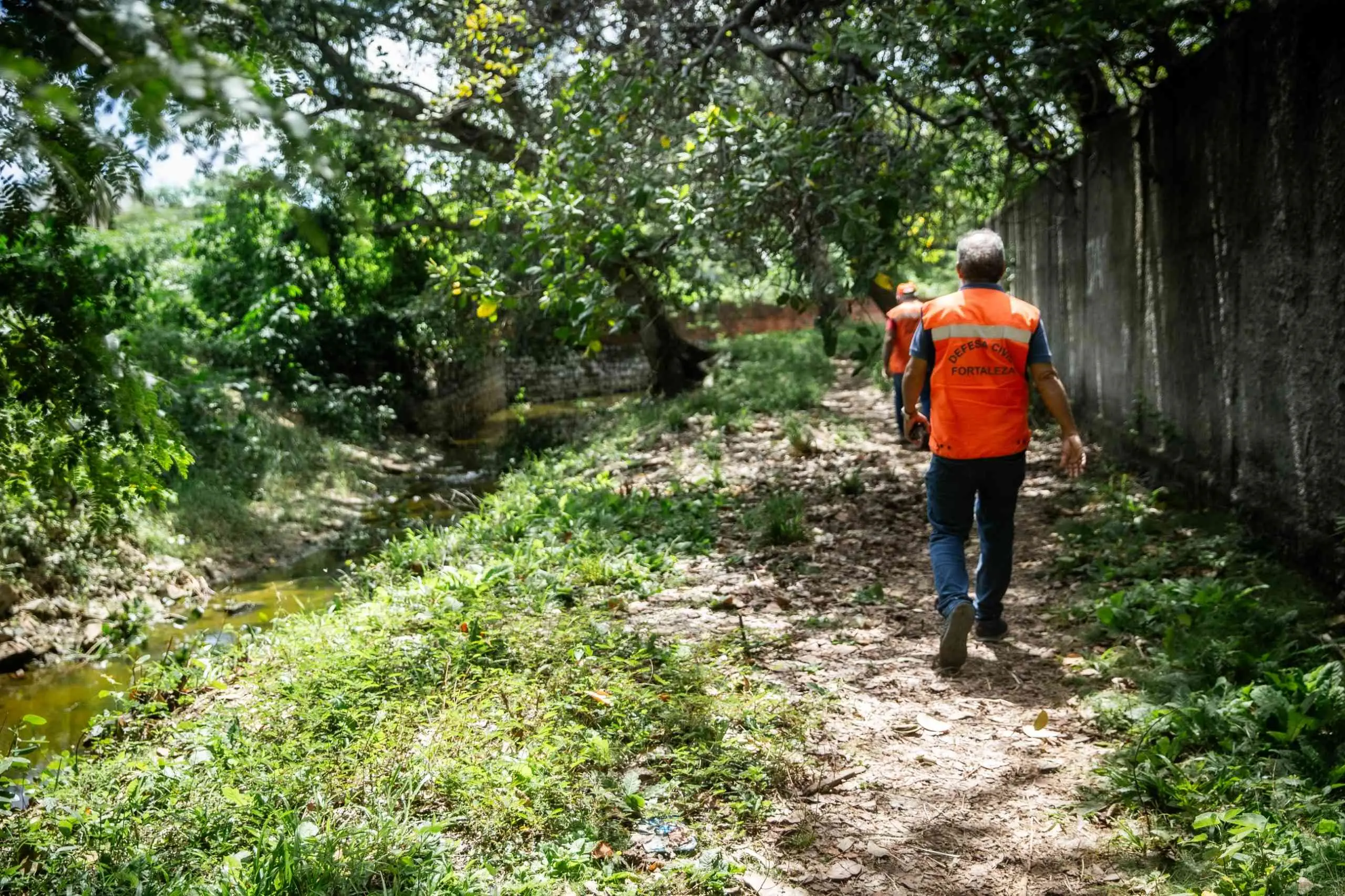 Agente da Defesa Civil de Fortaleza caminha por trilha arborizada ao lado de um curso d'água e um muro de concreto no bairro Jangurussu.