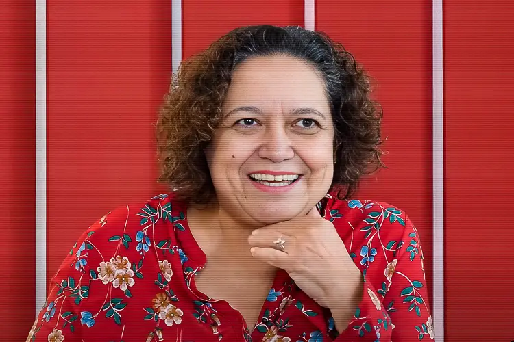 Sônia Braga sorri para a foto com a mão esquerda no queixo. Veste blusa florida com tom avermelhado.
